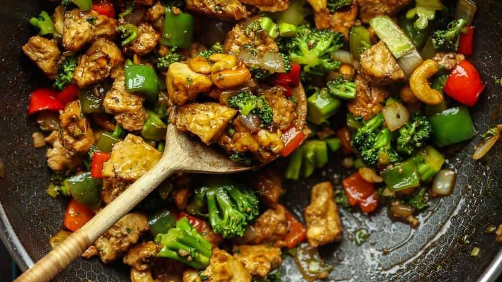 A wooden spoon holds a cashew nut stir fry with tofu, broccoli, green and red bell peppers, cashews, and onions in a pan. The dish is colorful and appears to be seasoned with a glossy sauce.